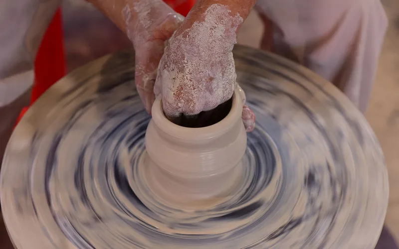 Les mains d'une femme façonnant de l'argile humide sur un tour de potier dans un atelier traditionnel en briques, lumière matinale filtrée