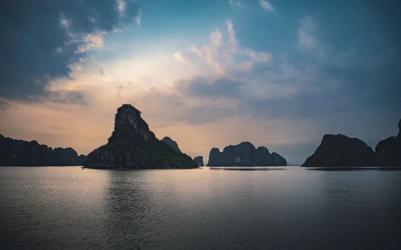 Wide aerial view of limestone karst islands rising from calm green water at golden hour, mist settling in the valleys between
