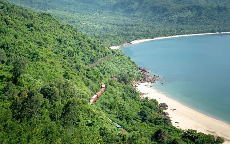 A quiet tropical beach at low tide, white sand curving into clear shallow water, dense green hills rising behind, late aftern