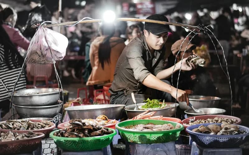 A Vietnamese coastal night market at dusk, warm lantern light over rows of seafood stalls, distant figures browsing, steam ri