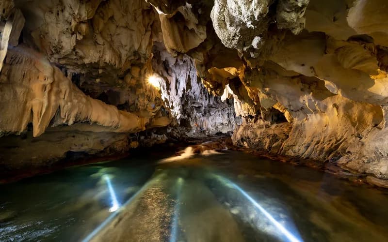 Intérieur d'une grande salle de grotte calcaire, un petit groupe de personnes en casques et gilets de sauvetage progressant dans une eau peu profonde