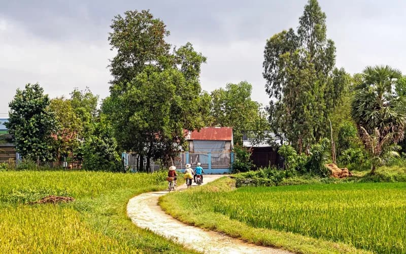 Un chemin de village vietnamien à l'heure dorée, un cycliste passant devant des maisons en bois avec des rizières de chaque côté, des pitons calcaires en arrière-plan