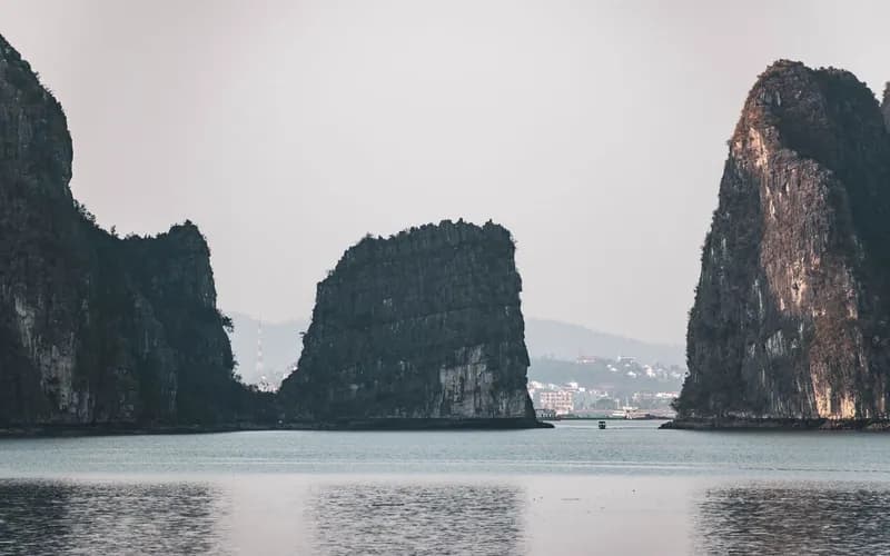 Vue large de la baie d'Ha Long à l'aube, karsts calcaires s'élevant d'une eau calme gris-vert, légère brume au ras de l'eau, petite embarcation en bois