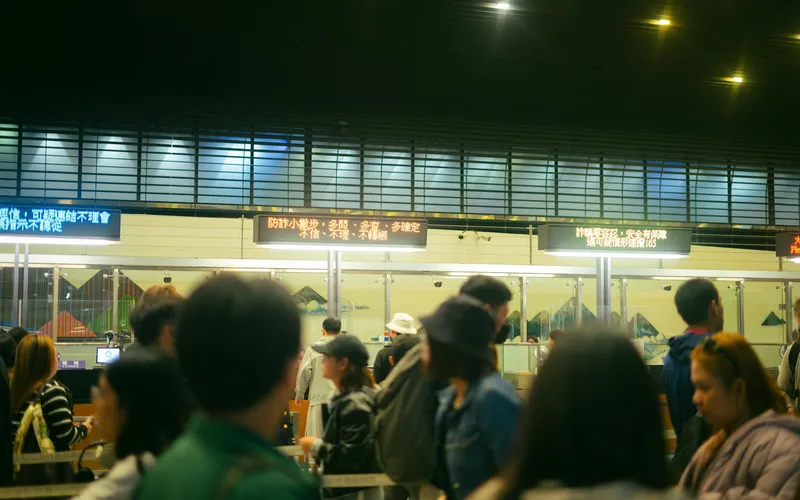 a traveler's hands holding a printed document near an airport immigration queue, overhead fluorescent lighting, other passeng