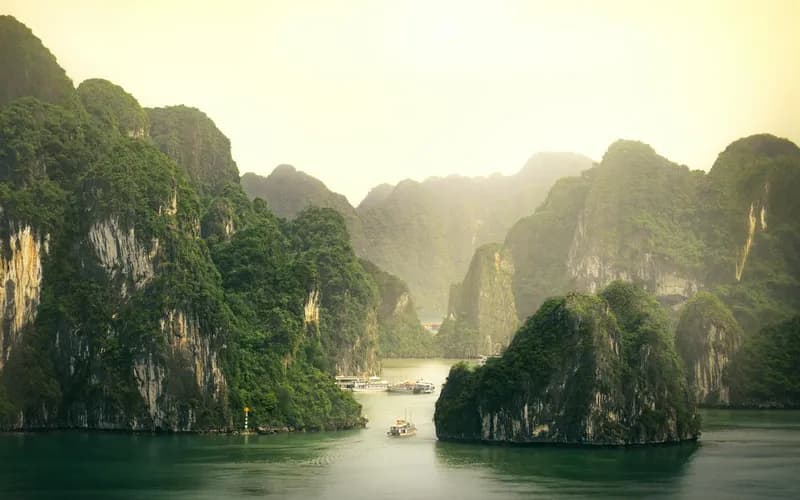 Un bateau en bois sur les eaux calmes de la baie d'Ha Long à l'aube, brume s'élevant entre les karsts calcaires, lumière dorée et chaude