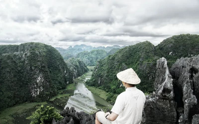 A lone traveler seated on a wooden platform overlooking a forested river valley in Vietnam, viewed from behind, late afternoo